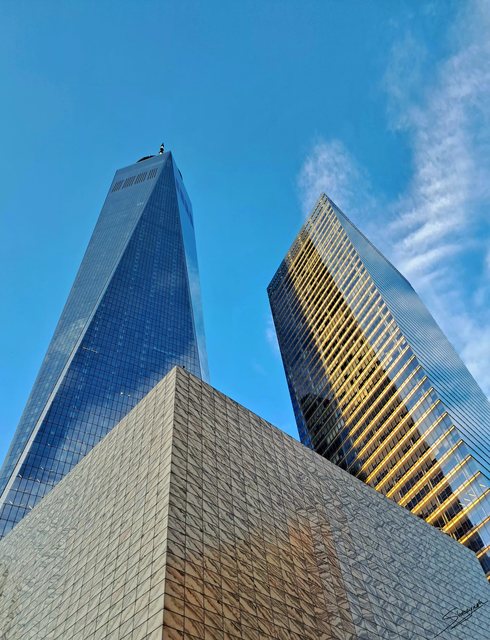 One World Trade Center and neighbouring buildings viewed from below against a blue sky.